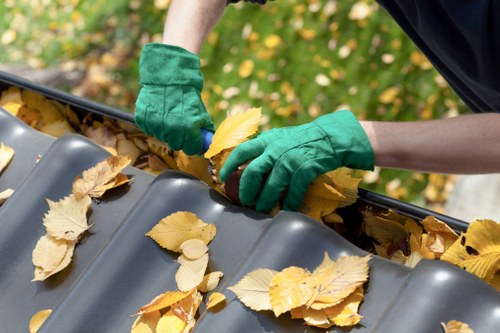 A person wearing green work gloves is collecting fallen yellow and brown leaves from a large, dark grey, corrugated plastic roof gutter. The gutter is partially filled with autumn leaves, which are scattered across its surface. The background shows a blurred outdoor environment with green foliage and sunlight filtering through trees, indicating an outdoor setting during autumn. The scene suggests routine maintenance as part of a waste removal or clearance service, possibly carried out by Commercial Waste Loughton, based on the visible context. The person's arm is visible, and they are carefully gathering the leaves without damaging the gutter, which appears clean apart from the leaves. The image highlights the importance of regular outdoor rubbish and waste removal to maintain property hygiene and prevent blockages in drainage systems, relevant to services offered in Loughton and surrounding areas within Essex.