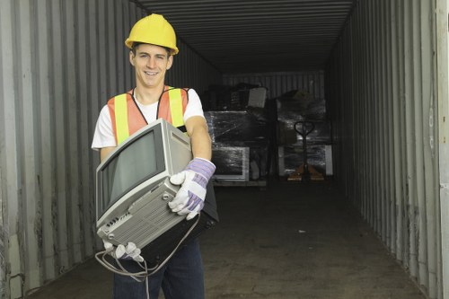 A young male worker wearing a yellow safety helmet, a high-visibility vest with reflective strips, a white t-shirt, and work gloves, is standing inside a metallic storage container or shipping space. He is holding an old, bulky computer monitor with a grey plastic casing and a curved screen, which appears to be discarded electronic waste. The background shows a dark interior with stacked black plastic-wrapped items and a pallet truck or trolley partially visible behind him, indicating an environment related to rubbish or waste clearance. The setting suggests a waste management or rubbish removal context, possibly within a commercial or industrial area, with the focus on proper disposal of electronic waste. The lighting is natural, and the scene emphasizes clean, organized waste collection activities in a location accessible from the surrounding environment, consistent with services offered by Commercial Waste Loughton in the local area.