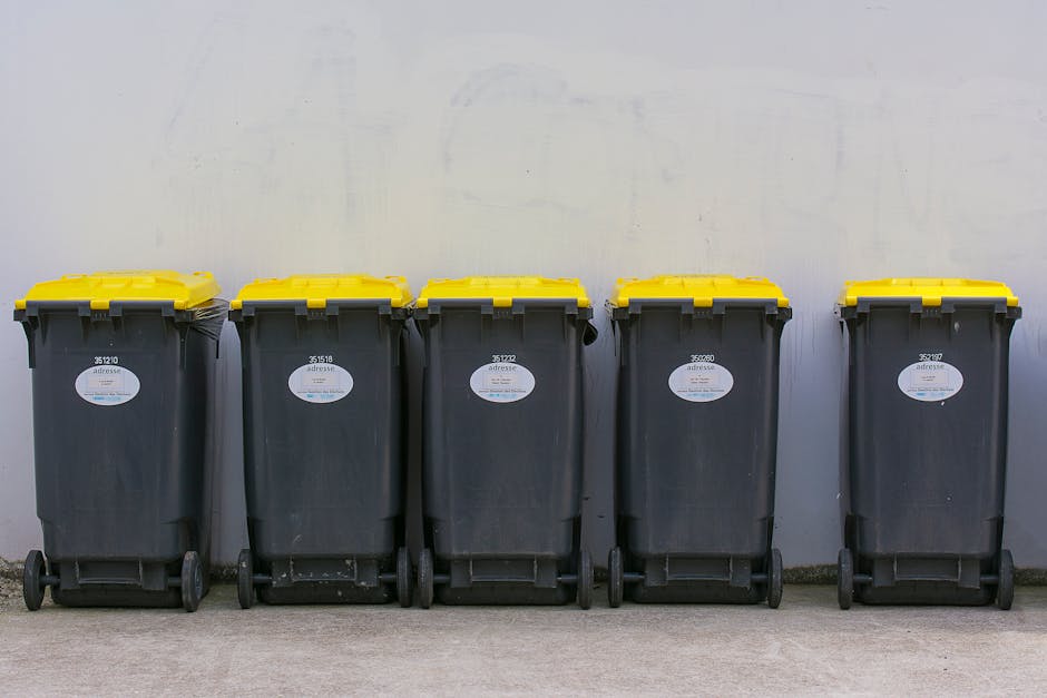 A waste collection worker wearing a yellow and red high-visibility vest stands at the rear of a large red rubbish truck, which is positioned on the side of a street. The truck's rear loading mechanism