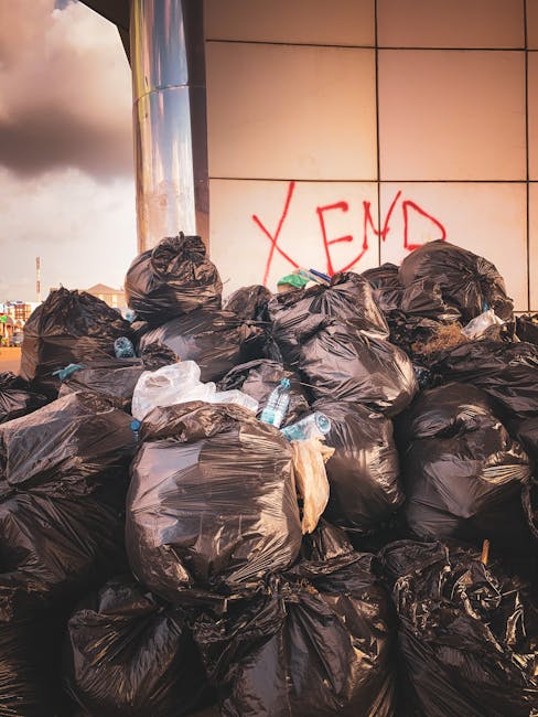 A large pile of black plastic rubbish bags stacked against a modern exterior wall composed of smooth, beige tiles with visible grout lines. The bags are tightly sealed and some appear slightly bulging